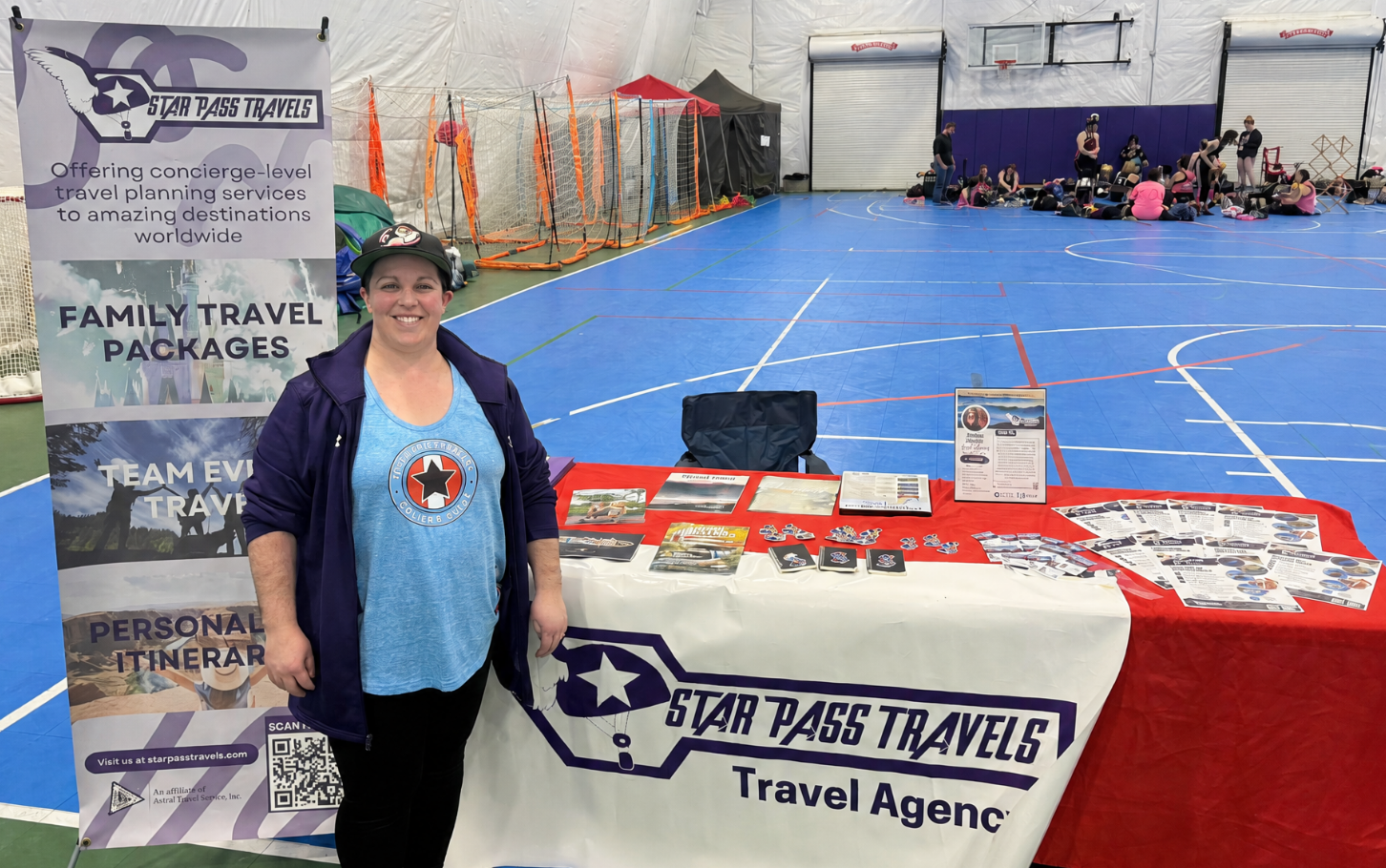 Photograph shows a travel agency booth inside an indoor sports facility, featuring Star Pass Travels signage, family travel packages, and personal traveler services. A smiling representative stands beside a branded table covered with brochures, cards, displays, and a QR code, while blue court, volleyball equipment, and recreational groups fill background.
